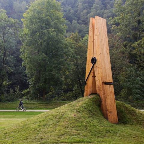A giant wooden clothespin sculpture by Mehmet Ali Uysal appears to pinch a grassy hill in a forested landscape. The playful design creates the illusion of the ground being pinched together.