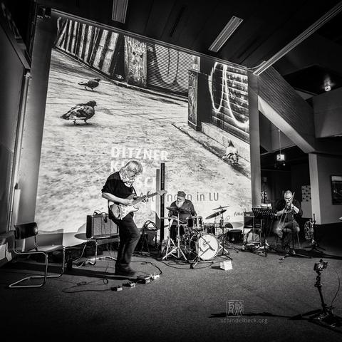 Schwarzweiß Fotografie von John King, Erwin Ditzner und Stephan Kirsch, die im Wilhelm Hack Museum in Ludwigshafen das Release Konzert für die CD "One Afternoon in LU", erschienen bei fixcel records, gespielt haben. Im Hintergrund das Cover der CD mit drei Tauben.