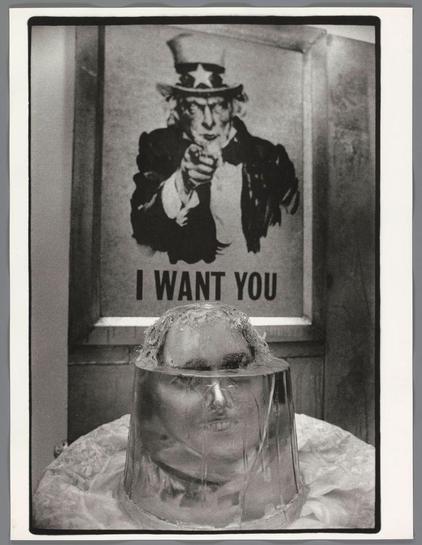 The image is a black and white photograph featuring two distinct subjects. In the foreground, there's an individual with their head encased in what appears to be a clear plastic covering or mask, partially obscuring their face. The person has visible strands of hair escaping from under the edge of this cover.

Behind them stands another figure wearing a traditional Uncle Sam hat—a tall top hat adorned with stars and ribbons—completing his iconic outfit which includes a suit jacket over a shirt and trousers. This character is pointing directly at the viewer, creating an imposing or directive gesture as if issuing a command or rallying someone to action.

Above this figurative image of Uncle Sam hangs a poster with bold text "I WANT YOU" prominently displayed across it in all caps. The style of both subjects suggests historical context but has been transformed into contemporary art through Kienholz's work, blending elements from the past with modern-day commentary or satire on identity and roles.

The photograph is set against what looks like an interior wall possibly within a building given its texture suggesting plasterwork or painted surfaces typical in indoor spaces. The juxtaposition of these two figures—Uncle Sam as a national symbol versus another obscured person—may provoke thoughts about individuality, society's expectations (as represented by Uncle Sam), and perhaps critique how individuals are sometimes perceive [...]