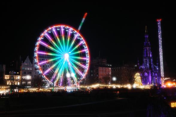 A view of the Edinburgh Christmas market during the night. A Ferris wheel, the Scott Monument, and other rides are lit up in decorations.