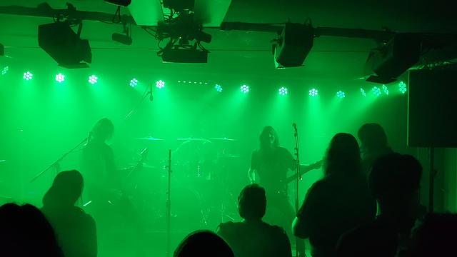 Die Band Zmey spielt bei grünem Bühnenlicht. Zu erkennen sind Gitarrist und Sänger, Bassist und ganz blass im grünem Licht verschwindend der Schlagzeuger. Die Bühnenhöhe ist sehr gering, das Rigging hängt an der Decke und das Publikum steht unmittelbar vor der Band.