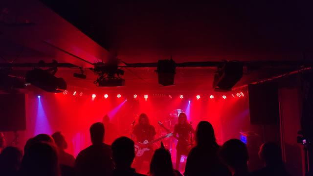 Es spielt die Band AstroKing auf einer rot beleuchteten Bühne. Zu sehen ist die Bassistin, der Sänger und Gitarrist, ein weiterer Gitarrist im roten Licht verschwindend. Den Schlagzeuger kann man leider nur erahnen. Die Bühnenhöhe ist sehr gering, das Rigging hängt an der Decke und das Publikum steht unmittelbar vor der Band. Es sind ca. 15 Köpfe aus dem Publikum unten im Bild zu sehen. Zu erkennen ist auch, dass mindestens eine dieser Köpfe ein spitzes Partyhütchen trägt.