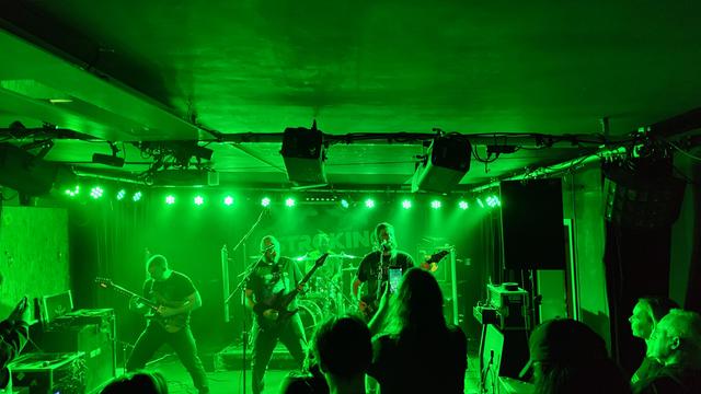 Es spielt die Band Countless Skies bei grünem Bühnenlicht. Das Backdrop ist von AstroKing, aber die Band auf dem Foto sind Countless Skies aus England. Zu erkennen  sind alle 4 Bandmitglieder. Die Bühnenhöhe ist sehr gering, das Rigging hängt an der Decke und das Publikum steht unmittelbar vor der Band. Auf der Bühne sieht man einiges an Equipment stehen, sowie eine große Lautsprecherbox auf der rechten Seite.