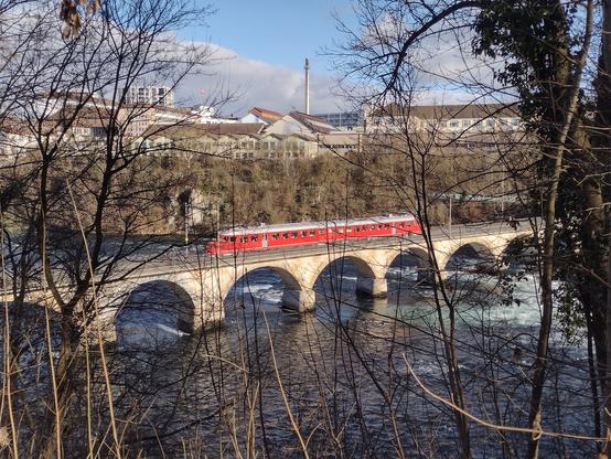 Ein roter alter SBB-Zug fährt über eine Steinbrücke (Brücke über den Rheinfall) mit mehreren Bögen. Die Brücke überspannt einen Fluss, dessen Wasser leicht bewegt erscheint. Im Hintergrund sind kahläumige Bäume und Gebäude zu sehen. Das Bild ist von Bäumen im Vordergrund leicht verdeckt, die im Winter kahl sind. Der Himmel ist hell und sonnig.