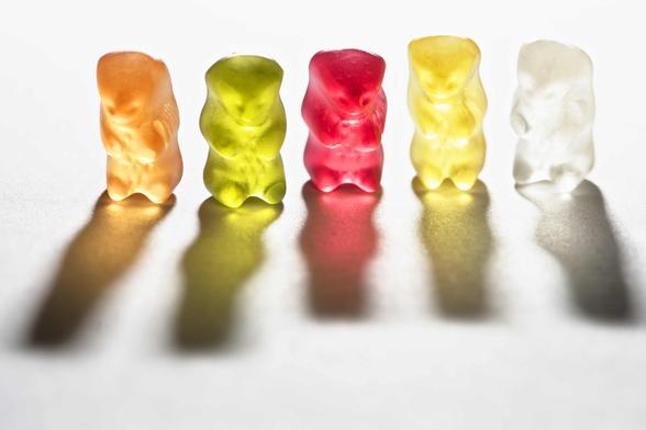 a number of gummibears lined up on a white sheet. They are backlit casting a colourful shadow.