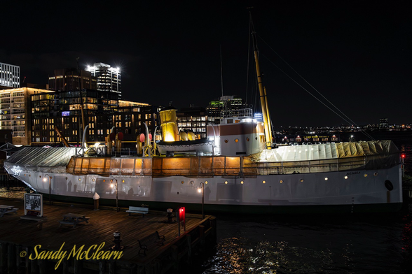 Old ship with winter tarps covering the deck at night with a cityscape in the background.