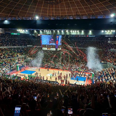Scene at the conclusion of a basketball game under the Big Dome of the Araneta Coliseum. White confetti and maroon-and-green streamers in the air, raining down on the brightly lit court, upon players and other people starting to gather at the center as one side has just won a championship. People have their phones out, capturing the precious, joyful moment.