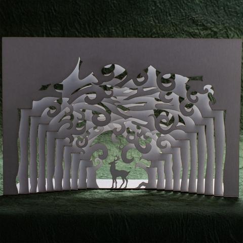 A pop up card made from cutting and folding a single sheet of stock depicts a stag standing framed by the convergence of rows of trees receding into the distance.  The branches overhead form whirls and spirals.  The background is a green textured paper (momigami) with the white card backlit against it.