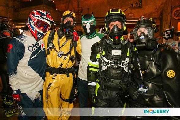 Group picture inside the Eloria main hall, with other gearheads and industrial walls in the background. On the left stands Focus571 in blue-white MX gear and a white-blue-red MX helmet with red MX goggles. Next to him the fox in his yellow Trellchem hazmat suit with full SCBA set including a gas mask, respirator and harness with air gauge hanging from his right shoulder and a belt. Next to the fox, in the middle of the picture, stands Treefox with a green-black-white puppy hood and a white-green T-shirt and jeans trousers. All three are facing the camera and hold each other in their arms.
On the right side of the picture stands Adensy, slightly in front of Treefox, wearing raingear with a MX chest protector, rubber gloves, MX neck protector, gas mask, helmet with ear protectors and an SCBA set with harness and respirator. To the right, arm in arm with Adensy, stands Exodiver in a black Viking rubber drysuit, rubber hood and an OTS full-face diving mask.
