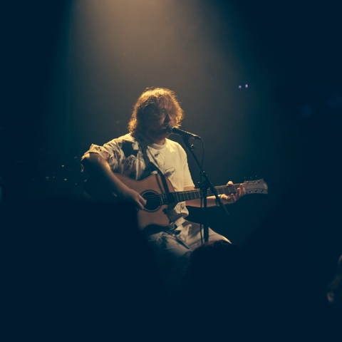 Feldup assis sur un tabouret, avec une guitare et un micro sur pied.