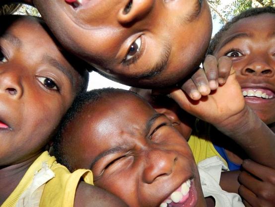 Five Malagasy children crowd together, pressing their faces close to the camera to fit into the frame. They are all making silly expressions, radiating energy and joy.