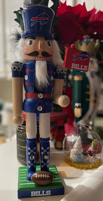 A tall blue nutcracker stands at the forefront off the image, with a smart royal blue jacket and boots, white pants, and a royal blue top hat.  His hat has a Buffalo Bills logo, he’s holding a red Buffalo Bills flag, and his stand is painted like a football field with a small football on it.  Go Bills!