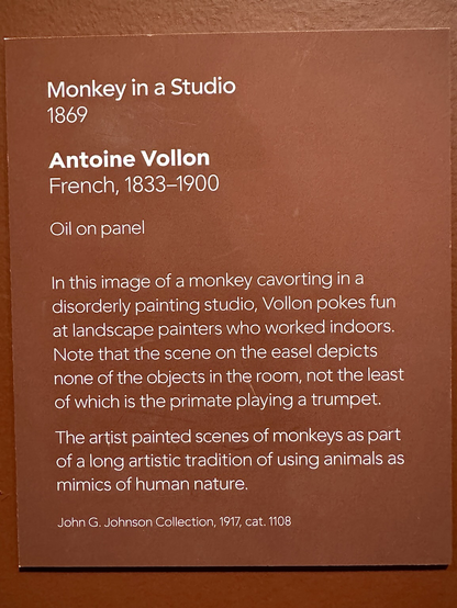 photo of rhe gallery label “In this image of a monkey cavorting in a disorderly painting studio, Vollon pokes fun at landscape painters who worked indoors. Note that the scene on the easel depicts none of the objects in the room, not the least of which is the primate playing a trumpet. The artist painted scenes of monkeys as part of a long artistic tradition of using animals as mimics of human nature.”