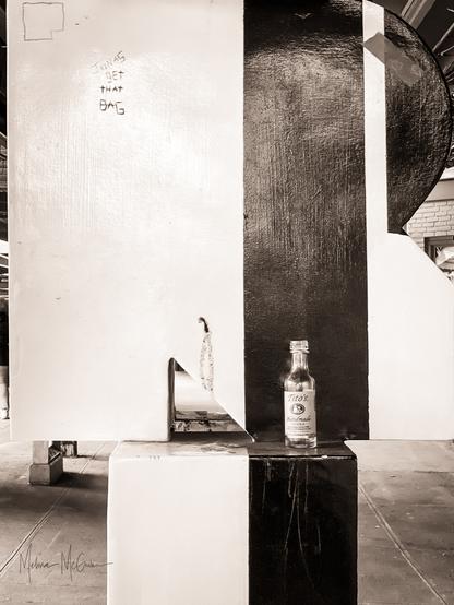 A black and white photo of a black and white sculpture at the New Rochelle Train Station. It's not obvious from the angle, but the sculpture is the letters N and R stacked on top of ones another, for New Rochelle. Someone has left behind on the sculpture a small, empty tequila bottle. At the top left of the sculpture, someone has scratched in the words, "Jonas, get that bag" into the surface.