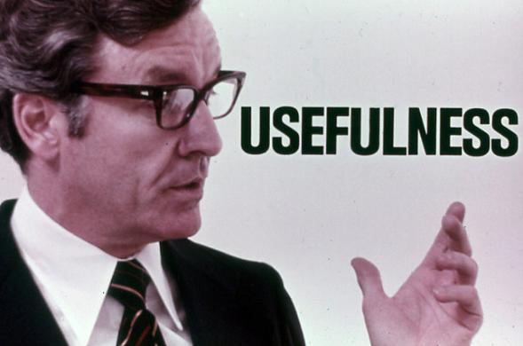 A man with a 1970s haircut and glasses and a black suit standing next to the word USEFULNESS