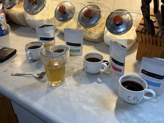 Four teacups filled with coffee surrounded by little coffee bags, a scale, a spoon, and a tall glass with rinsing water in it. There are lots of coffee drips on the counter.