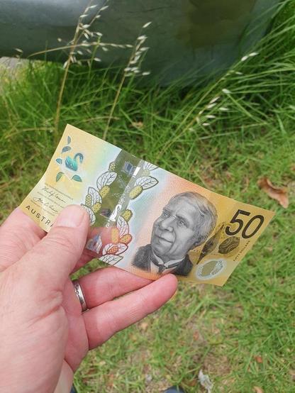 Closeup of an Australian $50 note held open in a hand, against a background of grass