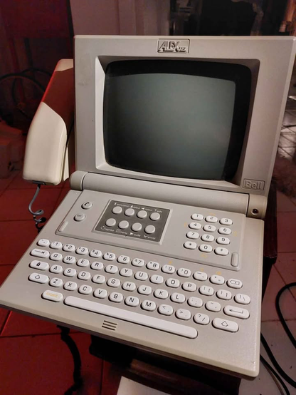 A gray videotex device comprised of a CRT, with a foldout keyboard with soft rounded keys. There's a telephone handset to the left.