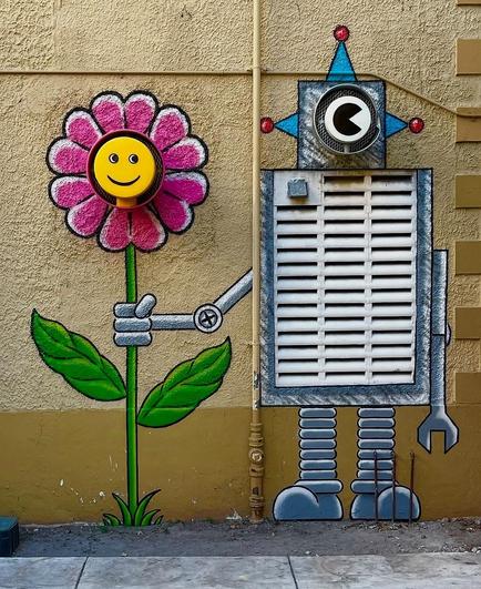 Streetart. A friendly robot with a flower was sprayed/painted on a wall with various parts of an air conditioning unit. The air conditioner was transformed into a robot and painted over. The rectangular grid is the belly of the robot, which has only one eye, which is a ventilation pipe from the wall. It has a star-shaped painted head, long legs and arms with round grippers. He stands upright and stretches one arm to the right towards a friendly smiling red flower that is just as big as he is. Another ventilation pipe with a round cover is the yellow smiley face of this flower. Very pretty and was painted for an elementary school.