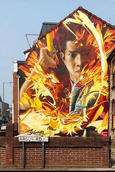 Streetartwall. A large mural of an Asian fighter is painted between flames on the outside wall of a two-storey furniture store. The face and one arm of a young man with short hair can be seen emerging from between an explosion of bright flames. The Wushu fighter is realistically depicted, and the conflagration fills the entire wall in bold orange/red colors with bright yellow arcs and peaks.
Info: Wushu means martial arts in Chinese and is often known as Kung Fu in the West. It is the generic term for all Chinese martial arts styles. The artist about the Mural: "This mural celebrates the dedication and spirit of a young man from Sibu who has mastered the ancient art of Wushu and risen to become a respected Sifu. Along the way, he embodies the profound teachings and disciplines of Wushu and demonstrates its grace, strength and philosophical depth."