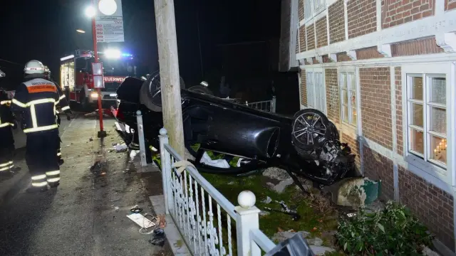 Ein dunkelgrauer oder schwarzer PKW liegt auf der Seite in einem Vorgarten vor einem Backsteingebäude. Das Auto ist erheblich beschädigt.  Feuerwehrleute in Schutzkleidung sind in der Nähe des Unfalls zu sehen. Ein Feuerwehrfahrzeug ist im Hintergrund sichtbar. Der Unfall ereignete sich nachts.