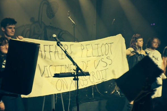 Tout le groupe de musique sur scène tenant une banderole avec d’écrit :
« Affaire Pelicot
victimes de VSS
on vous croit »
Tout en majuscule, le dernier mot est souligné.