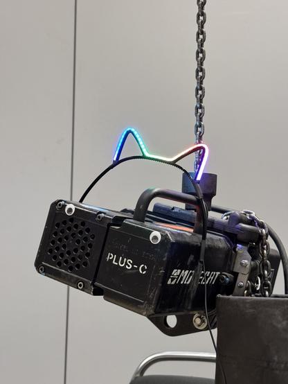 A photo of a motorised chain lift, normally used to lift and install stage equipment. „movecat“ is written in white letters on its side. It has glued on googly eyes and wears catears lit up in rainbow colours. It hangs on a chain that disappears at the top of the picture. The background is a white partition wall of an exhibition hall.
