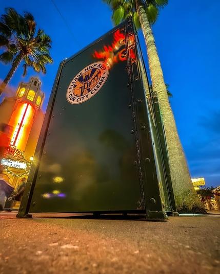 The image features a green trash can with a sign that reads "Please Help Keep Our City Clean." It is positioned in a city setting with palm trees and colorful lights in the background during twilight.