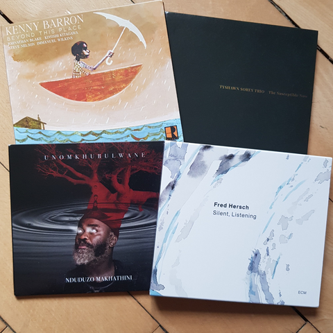assembly of CDs on wooden floor, clockwise: Kenny Barron "This Is The Place", Tyshawn Sorey Trio "The Susceptible Now", Fred Hersch "Silent, Listening", Nduduzo Makhathini "Unomkhubulwane".