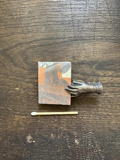 A decorative tiny silver hand-shaped clip holding a small book, positioned next to a matchstick on a wooden surface. The book features abstract designs in black and orange colors.