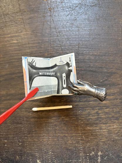 An intricately designed miniature book displaying a sewing machine illustration, held open by a decorative hand-shaped clip and red stamp tweezers.