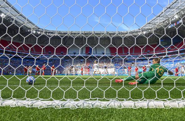 Blick von hinten durchs Tornetz auf den Elfmeter von Giulia Gwinn gegen Spaniens Torfrau Cata Coll. Coll hat sich auf die rechte Seite geworfen und blickt nach links dem Ball hinterher, der von Gwinn geschossen kurz davor ist, über die Linie zu rollen. Im Hintergrund ist zentral Gwinn nach ihrem Schuss, sowie alle anderen Spielerinnen, alle blicken auf den Ball, der Himmel über der Tribüne strahlt blau.