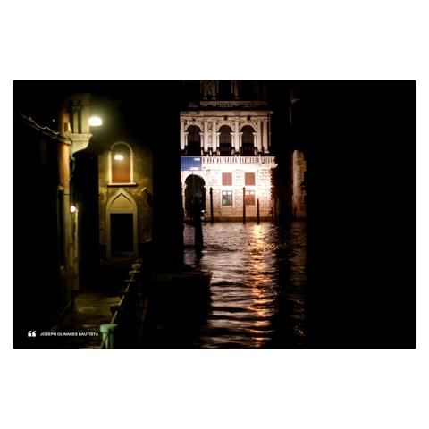 A nighttime scene in Venice, Italy, featuring a quiet canal with water reflecting golden light from illuminated buildings. A small walkway with glowing lamps leads toward the Grand Canal where a beautifully ornate building is bathed in light, highlighting its intricate architecture. Photo taken by Joseph Bautista.