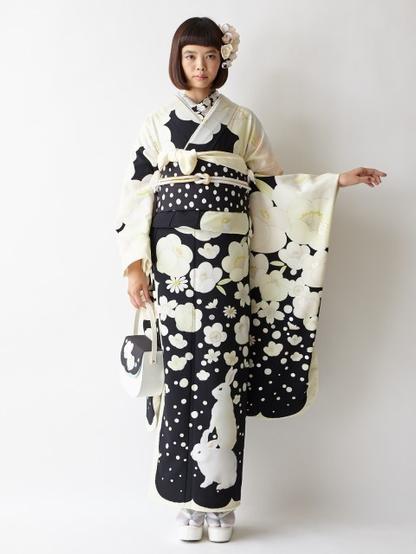 A feminine person with bobbed hair in front of a white background wearing a furisode kimono with a navy blue background within white rabbits at the bottom with white floral pattern above. The pattern is small at the bottom and grows larger towards the top. The person's left hand is held out to the side.