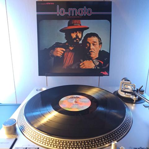 A vinyl record sits on a turntable. Behind the turntable, a vinyl album outer sleeve is displayed. The front cover shows Willie Colon holding a gun to Hector Lavoe's head.