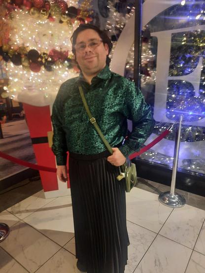 Me wearing a green and black silk shirt, a black skirt, with glasses, and wavy brown hair. Background is a Christmas tree.