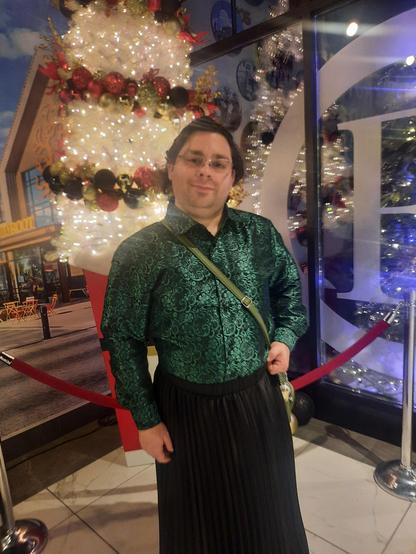 Me wearing a green and black silk shirt, a black skirt, with glasses, and wavy brown hair. Background is a Christmas tree.