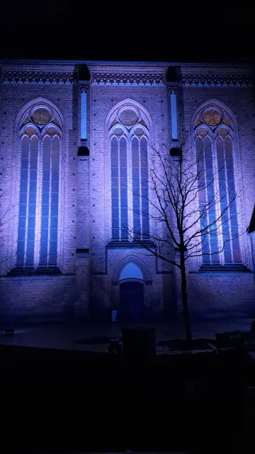 Foto - Anblick der Seitenfront mit großen Fenstern der Pfarrkirche in Backsteingotik in Güstrow, sie wird von unten hell angestrahlt, darüber der Himmel ist dunkel.