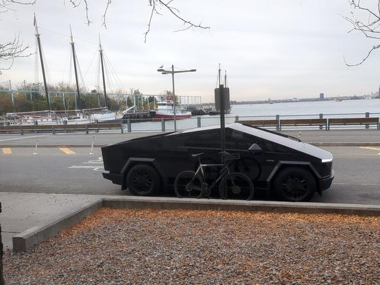 A black Tesla Cybertruck parked alongside the sidewalk in Brooklyn Bridge Park. Across the street we see the river, Brooklyn Bridge Park Pier 6, and two boats docked alongside Pier 6. There is a bike chained right in front of the Cybertruck.