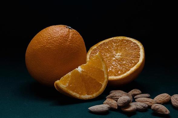 In this still life food photo, one whole orange, one half and an eighth slice are placed on a dark teal to black background, with some almonds scattered in the foreground.