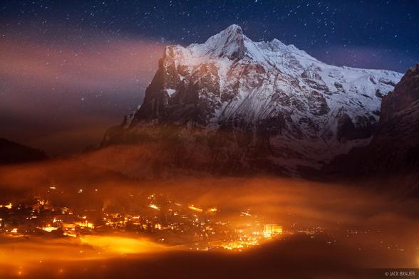 The lights of the town of Grindelwald shine in the fog, while the snowy mountain of Wetterhorn (3692m) towers above in the moonlight, December.