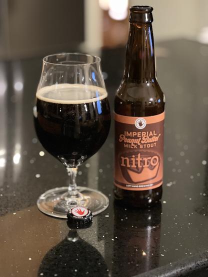 A tall tulip glass of dark beer next to a brown bottle with a label on shades of deep orange and brown. A stylized peanut butter cup has a crown shaped bite