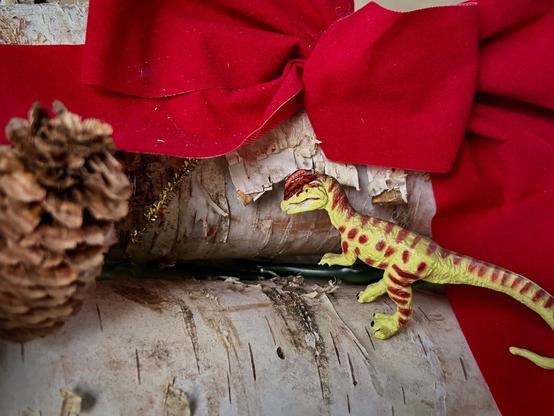 A green Dilophosaurus with brown spots stands on a stack of white birch logs decorated with a large red bow, pine cones and holly.
