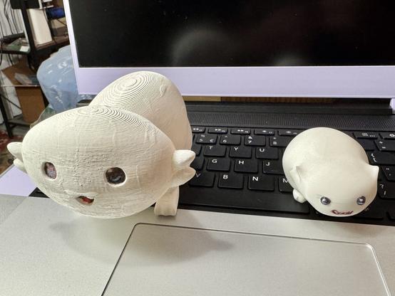 Larger and smaller 3D printed and hand-painted Moopsy sculptures sitting on a laptop keyboard. The larger of the two is on the left with a happy expression and the smaller has a scowling expression