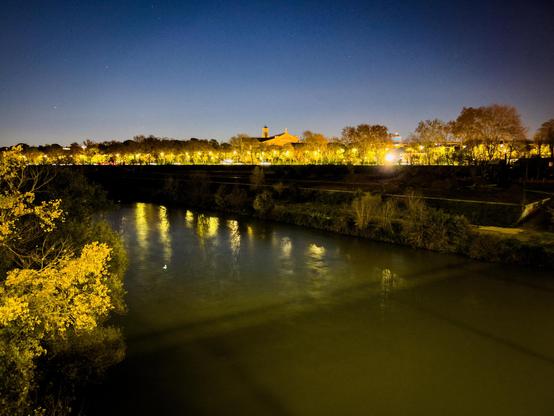 Scena notturna di un fiume con alberi lungo le sue rive. Sullo sfondo, ci sono edifici illuminati, probabilmente con lampioni che si riflettono sull'acqua. Il cielo è sereno e scuro, suggerendo una notte tranquilla.