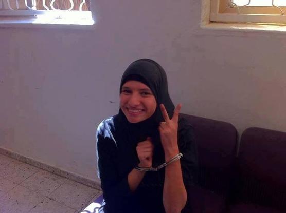 Das im Beitrag erwähnte Foto von Rouya Hzayel zeigt eine junge Frau mit Kopftuch in Handschellen, die vor einer Wand mit zwei vergitterten Fenstern auf einer violetten gepolsterten Bank  sitzt und mit einer Hand das Siegeszeichen macht.