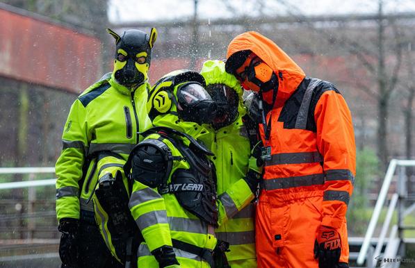 Before a blurred background of an industrial complex on a rainy day stand four gearheads under dripping water. In the middle stands Adensy looking up, in yellow hi-vis raingear and a Leatt MX chest and neck protector, gas mask and helmet with ear protectors with Exodiver right behind him, also in yellow hi-vis raingear, gas mask and with the hood up, while the water drips on them and sprinkles all around them. To the left, behind Adensy, stands Johnny the Buck in yellow hi-vis raingear and his black-yellow puppy mask, shaking off the water with his head. To the right next to Exodiver stands the fox in his orange hi-vis boilersuit with reflective stripes on the lower arms, belly and vertical stripes over the shoulders, orange-black puppy mask and with orange hood of the boilersuit on top. Exodiver and the fox look at the camera, with the fox having his head slightly tilted to the left.