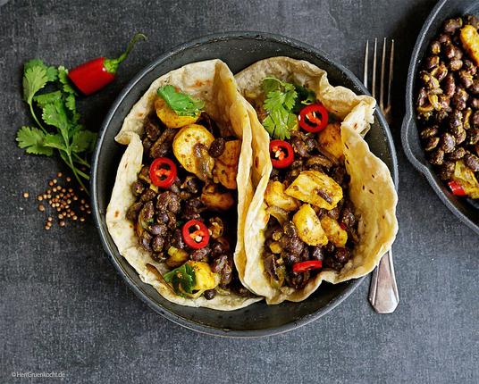 Dunkler Untergrund. Darauf eine schwarze Bowl. Darin zwei Tortillas mit Linsen, roten Peperonistücken und Bananenstücken.