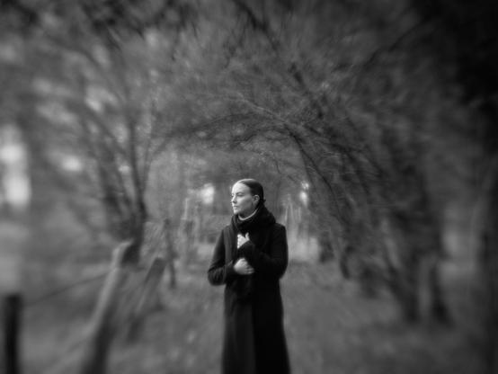 A young woman is standing on an overgrown dirt road at the edge of a forest and looking to the side, out of the picture. The image is quite blurred and has a wild bokeh.