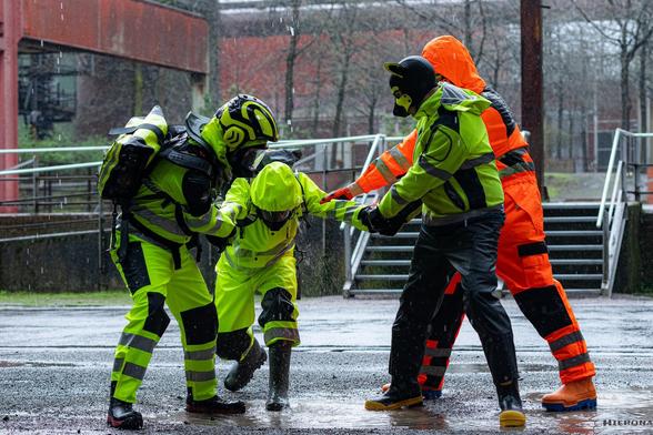 A rainy and grey day in an industrial complex. In the center is a water puddle on the ground and thick rain drips from the top. On the left stands Adensy in yellow hi-vis raingear and a Leatt MX chest and neck protector, gas mask and helmet with ear protectors and black rubber boots with red soles. To the right stands Johnny the Buck in a yellow hi-vis raingear jacket, black raingear pants, black rubber boots with yellow soles and his black-yellow puppy mask. Further to the right and behind Johnny the Buck stands the fox in his orange hi-vis boilersuit with reflective stripes on the lower arms, belly and vertical stripes over the shoulders, orange rubber boots with blue soles, his orange-black puppy mask and the orange hood of the boilersuit on top. They all are looking down to the center of the picture where Exodiver stands bent over, in his yellow hi-vis raingear and gas mask with raingear hood over his head and dark green rubber boots. All three hold Exodiver by his arms and try to pull him into the water puddle, while rain drips splatter on his back.